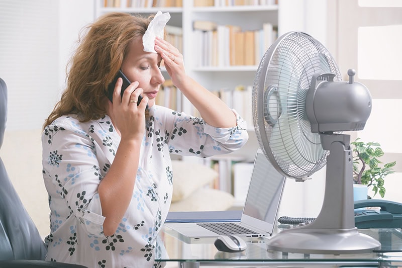 When Do I Replace My Air Conditioner? Woman sweating in front of a fan, while wiping the sweat off her forehead.