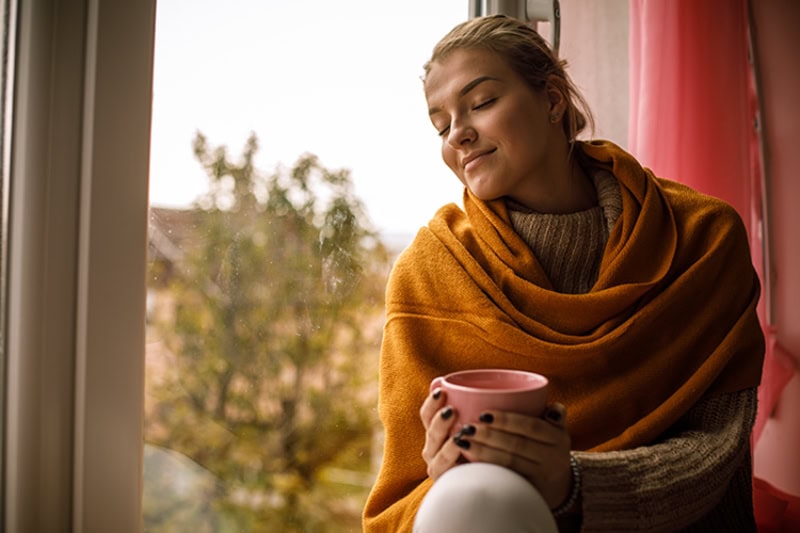 5 Tips to Improve Your Indoor Air Quality This Fall. Photo of a woman enjoying a fall view by a window inside of her home, drinking a hot beverage.